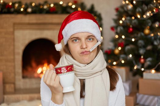 Young ill woman in santa hat, wrapped in scarf, measuring temperature and holding cup of warming drink, looking at camera, being sick on Christmas holiday, posing in room with new Year decorations.
