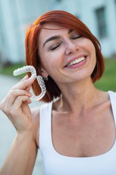 Red-haired Caucasian woman holding transparent mouthguards for bite correction outdoors. A girl with a beautiful snow-white smile uses silicone braces.