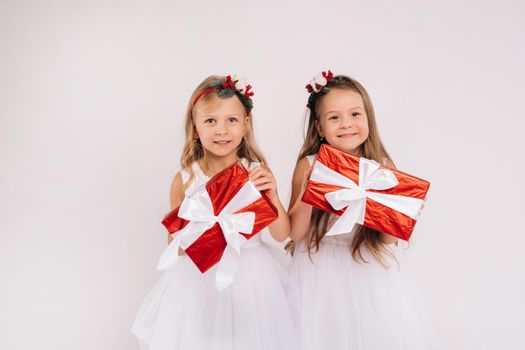 two little girls in white dresses with Christmas gifts on a white background smile.