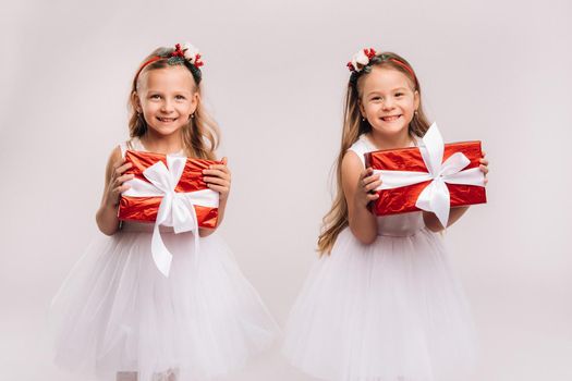 two little girls in white dresses with Christmas gifts on a white background smile.