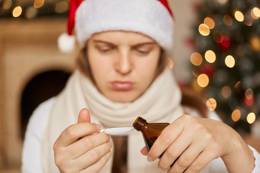 Sick on New Year! Sad ill woman in santa hat and white scarf sitting with medication in hands, needs to drink cough syrup, pouring it to spoon, female on blurred background.