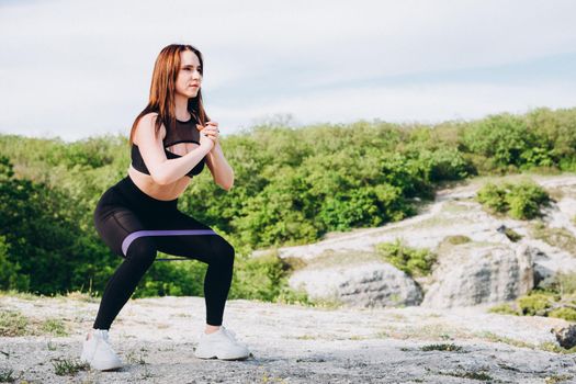 A beautiful athlete in the mountains. The girl is engaged in sports in nature. Purple rubber band for stretching and swaping muscles of hands and feet.