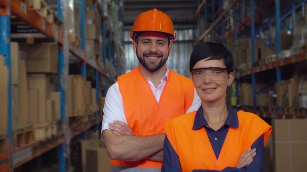 Portrait two workers in warehouse. Happy smiling professional Staff posing looking at the camera with friendly smile in logistic center. Young woman and handsome man wearing uniform high visibility orange hard hat and vest