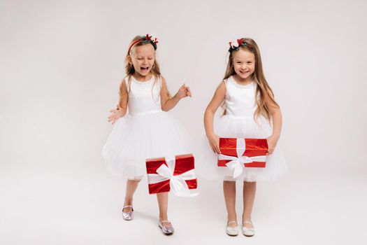 two little girls in white dresses with Christmas gifts on a white background smile.