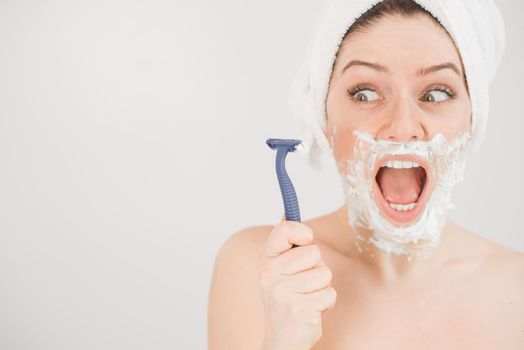 Funny portrait of a woman with shaving foam on her face holding a razor on a white background. The girl removes the mustache and beard.