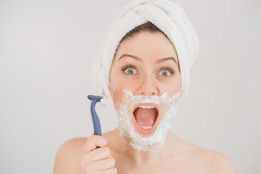 Funny portrait of a woman with shaving foam on her face holding a razor on a white background. The girl removes the mustache and beard.