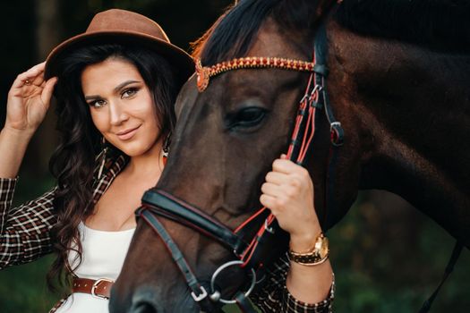 pregnant girl with a big belly in a hat next to horses in the forest in nature.Stylish girl in white clothes and a brown jacket