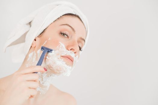 Cheerful caucasian woman with a towel on her head and shaving foam on her face holds a razor on a white background.