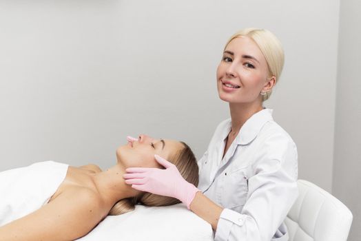 Beautician in rubber gloves gives the client an anti-aging massage in a beauty salon, looking at the camera.