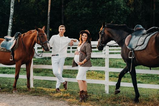 a pregnant girl in a hat and a man in white clothes stand next to horses near a white fence.Stylish pregnant woman with a man with horses.Married couple