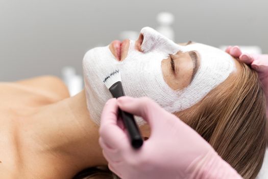 Beautician applies a white clay face mask with a brush on a woman's face. Professional beautician applies white cosmetic cream to a girl lying on a couch in a modern spa. Close up. Relaxed woman enjoying wellness facials.