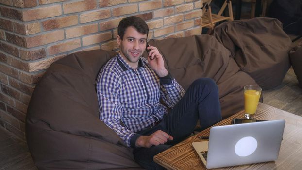 A man in a plaid shirt sitting in a chair holding a phone in his hand, the man talking on the phone on the table is a laptop and documents, looking at the camera.