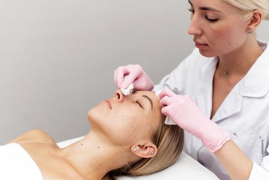 Close up of beautician's hands doing facial cleansing of young woman using cotton pads or sponge in beauty clinic or spa. Cosmetological treatment. Perfect skin concept. Young beautician or dermatologist cleans face of lying woman with cotton pads in spa salon.