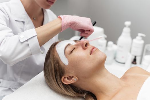 Beautician applies a white clay face mask with a brush on a woman's face. Professional beautician applies white cosmetic cream to a girl lying on a couch in a modern spa. Close up. Relaxed woman enjoying wellness facials.