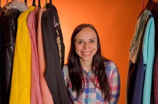 Young woman grimacing standing between clothes in wardrobe. Adult female choosing things in store