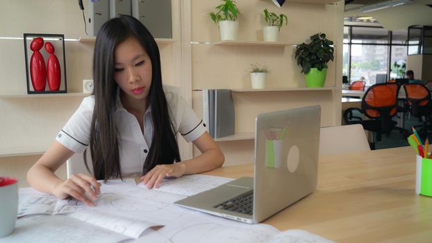 Female looking on blueprint and checking with electronic version on computer. Asian young professional designer works at the working place with table laptop . Multicultural smiling worker wearing in casual white shirt and black trousers