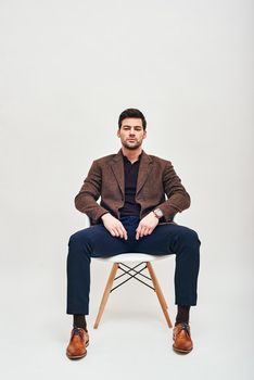 Full-length portrait of stylish dark-haired man wearing blue trousers and brown jacket, sitting on a white chair, leaning back and looking at camera isolated over white background. Front view
