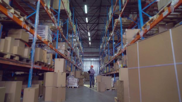 Man workers warehouse going along racks. Manager wearing hard hat writing notes in paper documents checking stock. Young employee with beard wearing casual shirt and jeans.