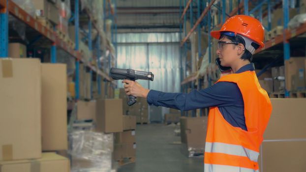 Young woman trainee working at warehouse. Worker wearing wearing high visibility clothing and a hard hat talking by phone and scans labels on boxes