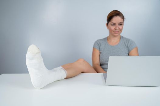 Caucasian woman lifted her leg with plaster to work desk and works on laptop on white background.