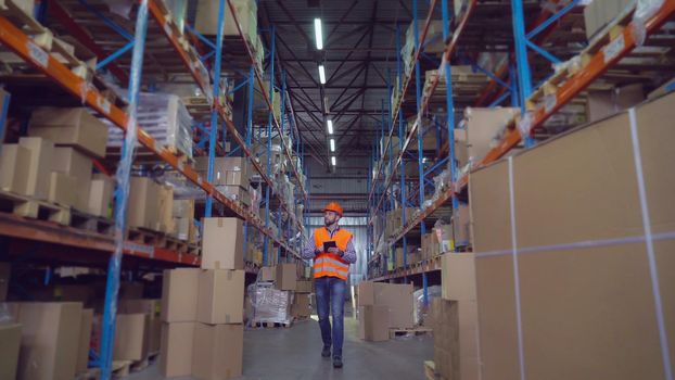 Man worker at warehouse going along metal shelving with boxes holding digital tablet. Manager at storehouse entering data for logistic.