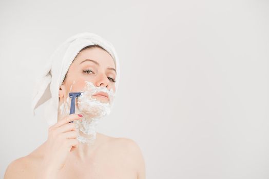 Cheerful caucasian woman with a towel on her head and shaving foam on her face holds a razor on a white background.