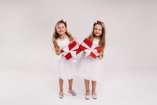 two little girls in white dresses with Christmas gifts on a white background smile.