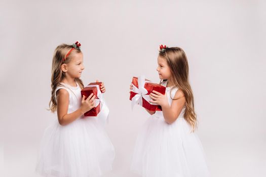 two little girls in white dresses with Christmas gifts on a white background smile.