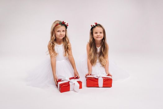 two little girls in white dresses with Christmas gifts on a white background smile.