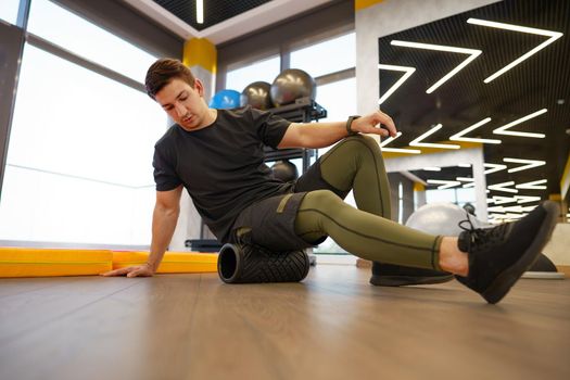 The man is massaging his leg muscles with the foam roller in gym