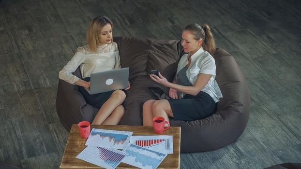 Two beautiful business woman in skirts resting on the couch, holding a laptop girl, the other girl talking on the phone. On the coffee table there are two cups of tea. Soft ponorama.