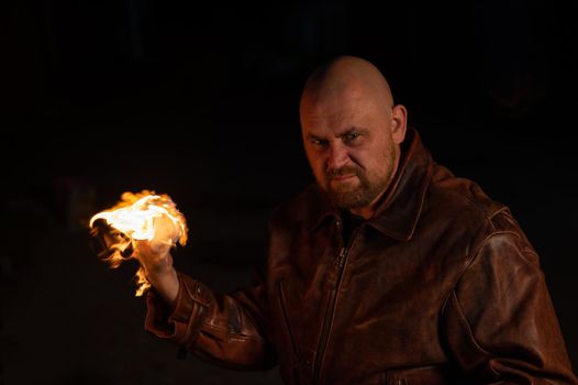 A bald man in a leather jacket holds a burning ball in his hand