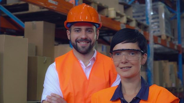 Portrait two workers at warehouse. Happy smiling co workers posing looking at the camera and smiling. Young professional woman and man wearing uniform high visibility orange hard hat ,vest