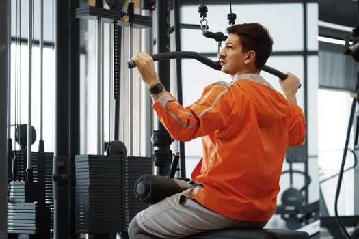 Young man in orange windbreaker working out training back muscles in the gym