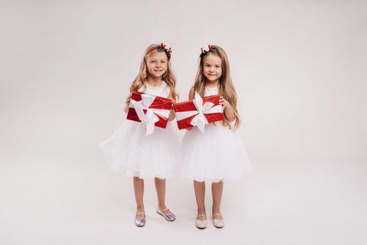 two little girls in white dresses with Christmas gifts on a white background smile.