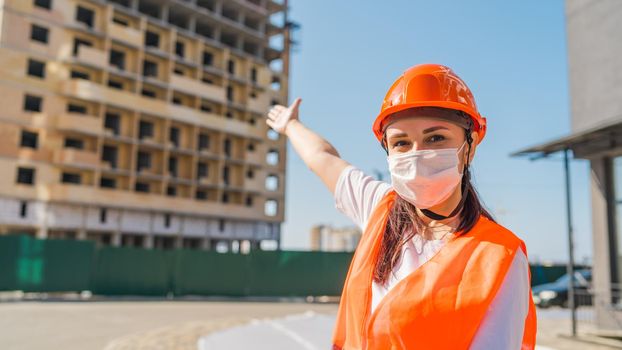 Female construction worker in overalls and in medical mask pointing at house under construction