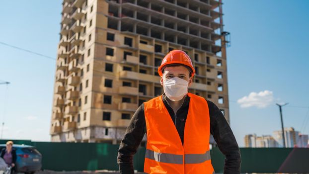 Portrait of male construction worker in medical mask and overalls on background of house under construction