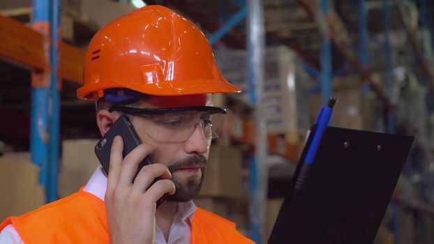 Portrait manager at work in warehouse. Handsome worker talking by smartphone discussing the logistics . Man with beard wearing hard hat and orange vest holding documents and checking delivery.