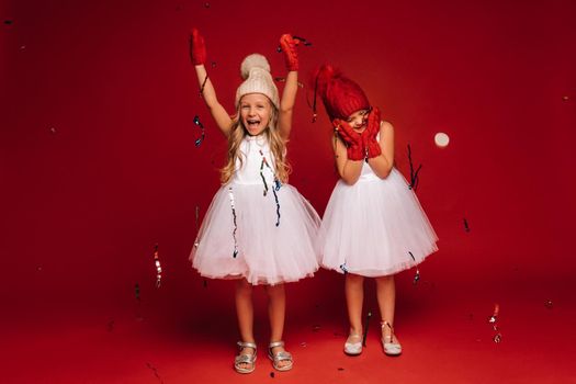 two little girls in white dresses hats and mittens sprinkle confetti on a red background.