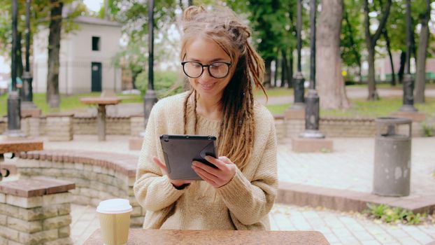 cheerful young caucasian woman with dreadlocks has coffee break in city using digital tablet scrolling touch screen