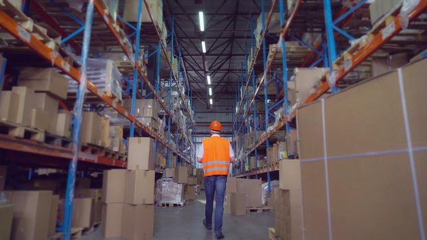 Manager wearing hard hat, safety vest and white shirt using digital tablet checking stock. Rear back view. Male workers warehouse going along metal shelves .