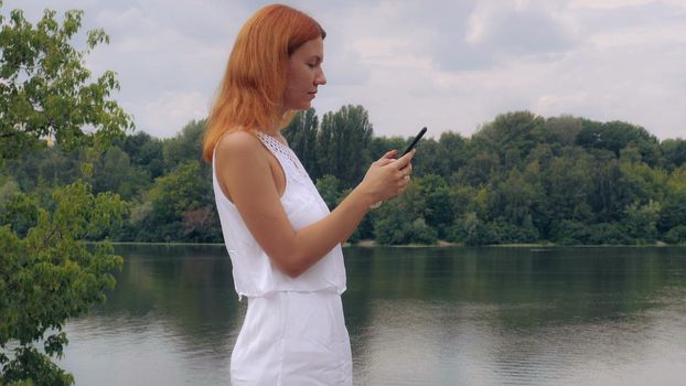 Young beautiful woman with red long hair standing near river or lake using smart phone. Attractive girl messaging or send email outdoors in summer season.