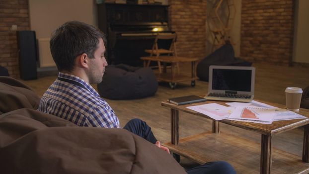 Attractive man sitting on the couch waiting for a meeting with a partner. On the table laid out charts and tables, next to a laptop lies diary.