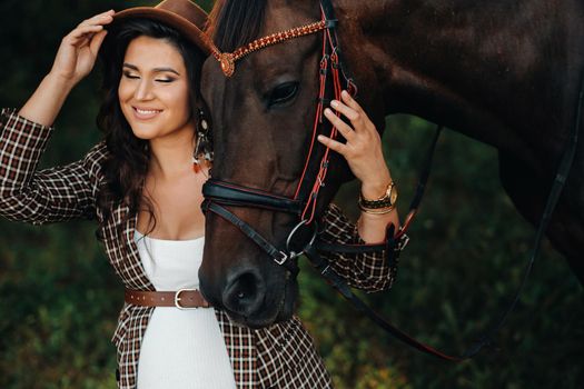 pregnant girl with a big belly in a hat next to horses in the forest in nature.Stylish girl in white clothes and a brown jacket