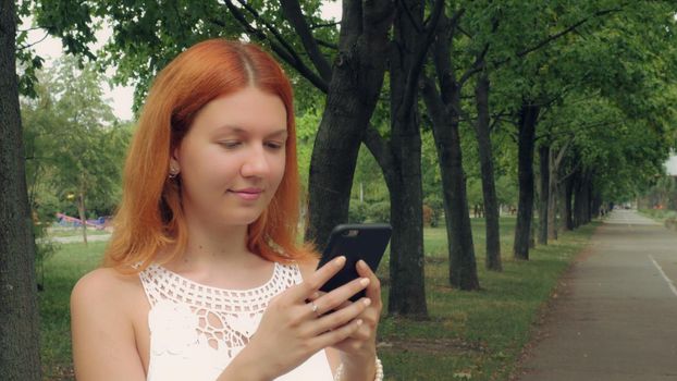 Redheaded woman texting on mobile phone in city with green nature summer season outdoors. Beautiful millennial girl using internet send email or sharing in social media or typing message on touch screen smartphone.