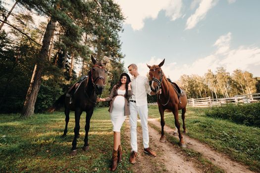 a pregnant girl in a hat and her husband in white clothes stand next to horses in the forest in nature.Stylish pregnant woman with a man with horses.Family