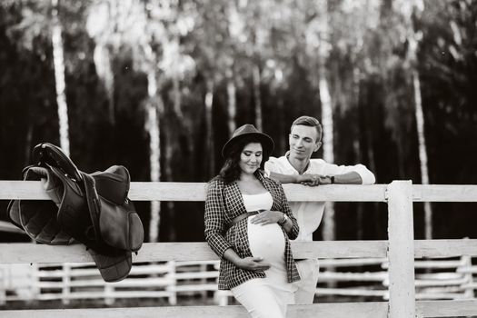 a pregnant girl in a hat and her husband in white clothes stand next to a horse corral at sunset.a stylish couple is waiting for a child in nature. black and white photo.