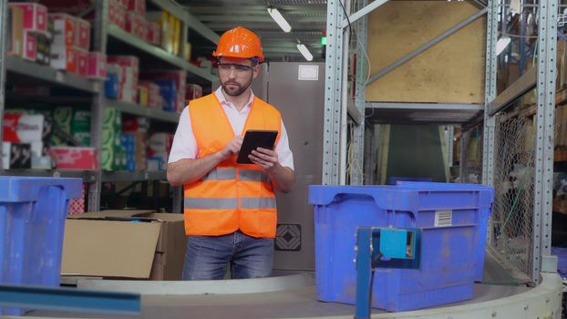 Warehouse worker standing near conveyor with parcels. Manager using digital tablet entering data. Man controls logistic wearing uniform and white shirt.