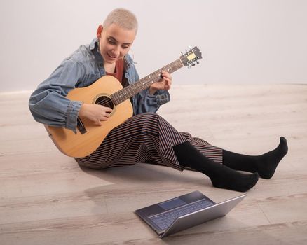 Young hipster woman sitting on the floor at home and watching guitar tutorials on laptop. Distance learning music in quarantine.