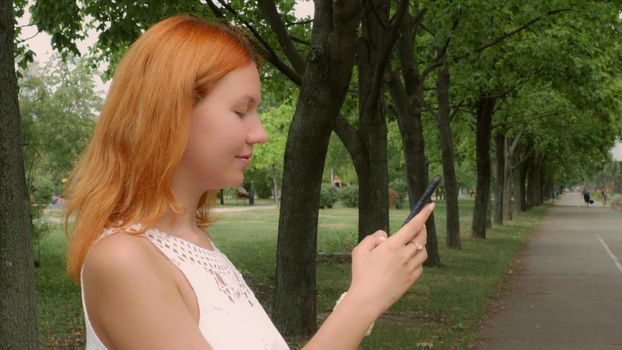 Young ginger student standing on the street in park or city using smart phone messaging. Beautiful millennial girl using wifi send email or sharing in social media or typing message on touch screen smartphone.